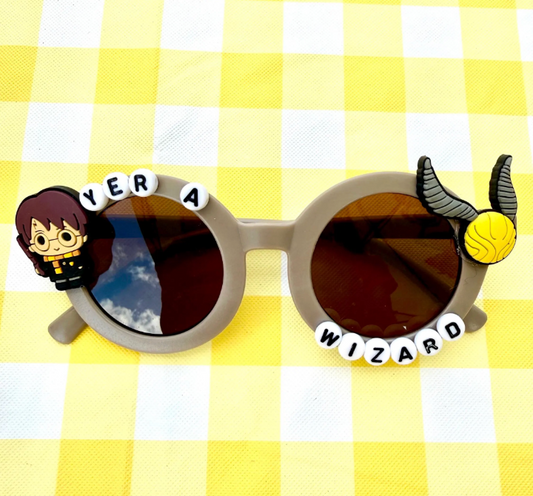 Kids grey round sunglasses with Harry Potter charm on left and Snitch charm on right with "Yer a Wizard" text around the frames.